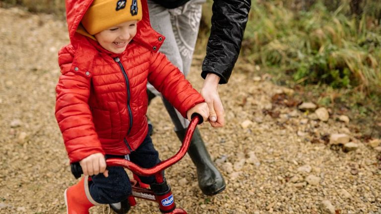 Joyful child riding a bike with parent on a gravel path, enjoying family time outdoors.